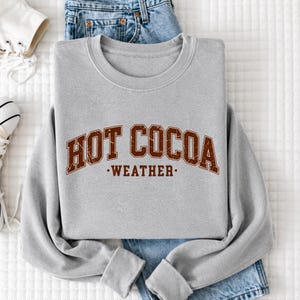 A gray sweatshirt with the text "HOT COCOA WEATHER" printed on it, along with a pair of blue jeans and a white sneaker.