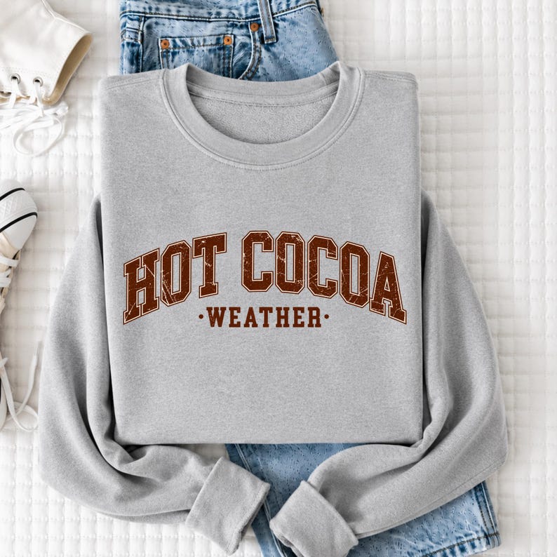 A gray sweatshirt with the text "HOT COCOA WEATHER" printed on it, along with a pair of blue jeans and a white sneaker.