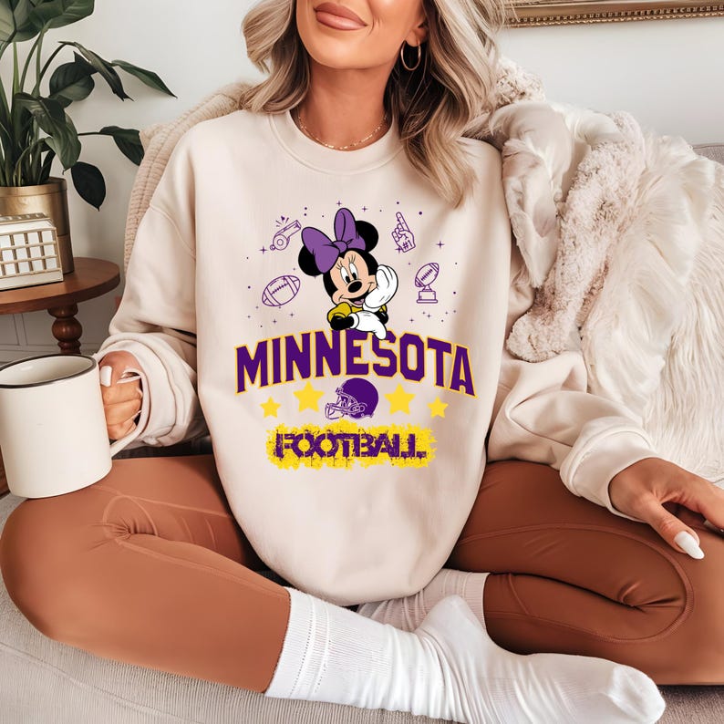 May include: Cream-colored sweatshirt featuring a Minnie Mouse graphic with a purple bow, holding a football. The word "MINNESOTA" is printed in purple, with yellow stars and the word "FOOTBALL" below. The sweatshirt is worn by a person.