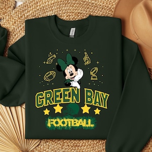 May include: Dark green sweatshirt featuring Minnie Mouse with a green bow, surrounded by football-themed graphics. The words "GREEN BAY FOOTBALL" are displayed in yellow and green, with stars and a football helmet graphic.