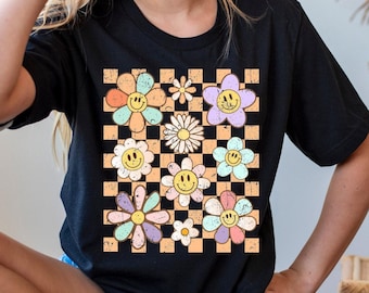 May include: A black short-sleeve t-shirt featuring a central graphic print of a tan and black checkered pattern overlaid with various colorful, distressed-style flowers. Several of the flowers have yellow smiley faces in their centers.