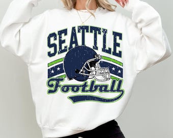 May include: A white long-sleeve sweatshirt featuring a distressed graphic print with the text SEATTLE Football in navy blue and lime green. The design includes a navy blue football helmet illustration centred between the text elements.