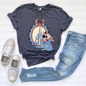 May include: A dark gray t-shirt featuring a Minnie Mouse graphic in a blue dress, with a castle backdrop. The graphic is framed by a pink bow and a blue oval border. The shirt is paired with light blue jeans and silver sneakers.
