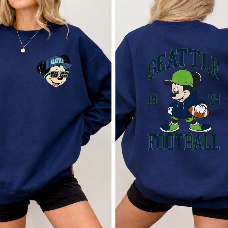 Puede incluir: Sudadera azul marino con un dise&ntilde;o de Mickey Mouse. La parte delantera muestra a Mickey con gafas de sol y un sombrero con "SEATTLE". La parte trasera muestra a Mickey con un bal&oacute;n de f&uacute;tbol, el texto "SEATTLE FOOTBALL" y los n&uacute;meros "19" y "74".