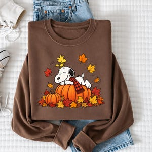 May include: A brown crewneck sweatshirt featuring a graphic of a white dog with a red and black plaid scarf lying on a large orange pumpkin. A small yellow bird sits on the dog's head, surrounded by scattered red, orange, and yellow autumn leaves.