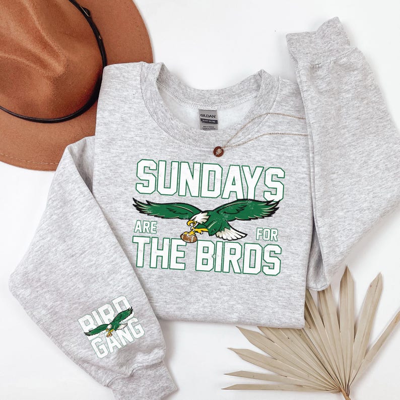 May include: A light gray sweatshirt with the text "SUNDAYS ARE FOR THE BIRDS" in green and white, along with a green and white eagle graphic. The sleeve features the text "BIRD GANG" and an eagle graphic.