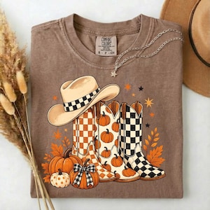 May include: A brown t-shirt featuring a graphic of a beige cowboy hat and two cowboy boots with checkered patterns. The design includes orange pumpkins, autumn leaves, and small stars. The shirt has a Comfort Colors brand label at the neck.