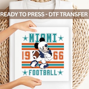 May include: A white DTF transfer featuring a cartoon character in a football uniform. The design includes the text "MIAMI", "1966", and "FOOTBALL" in teal and orange. The transfer is held up by hands.