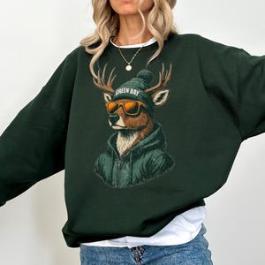 May include: A dark forest green crewneck sweatshirt featuring a graphic illustration of a deer wearing a dark green beanie with the text GREEN BAY, orange-tinted sunglasses, and a matching dark green zip-up hoodie layered over its fur.