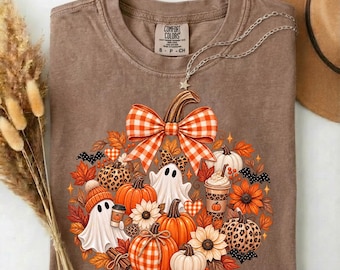Può includere: Una t-shirt marrone a maniche corte con una stampa grafica di zucche, fantasmi, girasoli e un grande fiocco a quadri arancione e bianco. Il design include dettagli leopardati, pipistrelli e una tazza di caffè. La maglia ha un'etichetta.