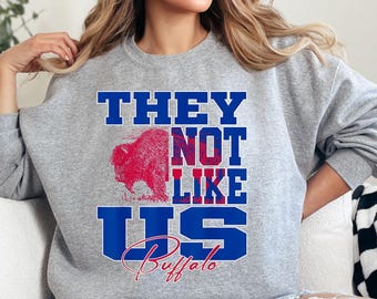 May include: A heather gray crewneck sweatshirt featuring a graphic print in blue and red. The text reads "THEY NOT LIKE US" in bold blue block letters, with a red buffalo illustration and the word "Buffalo" in a red script font below.