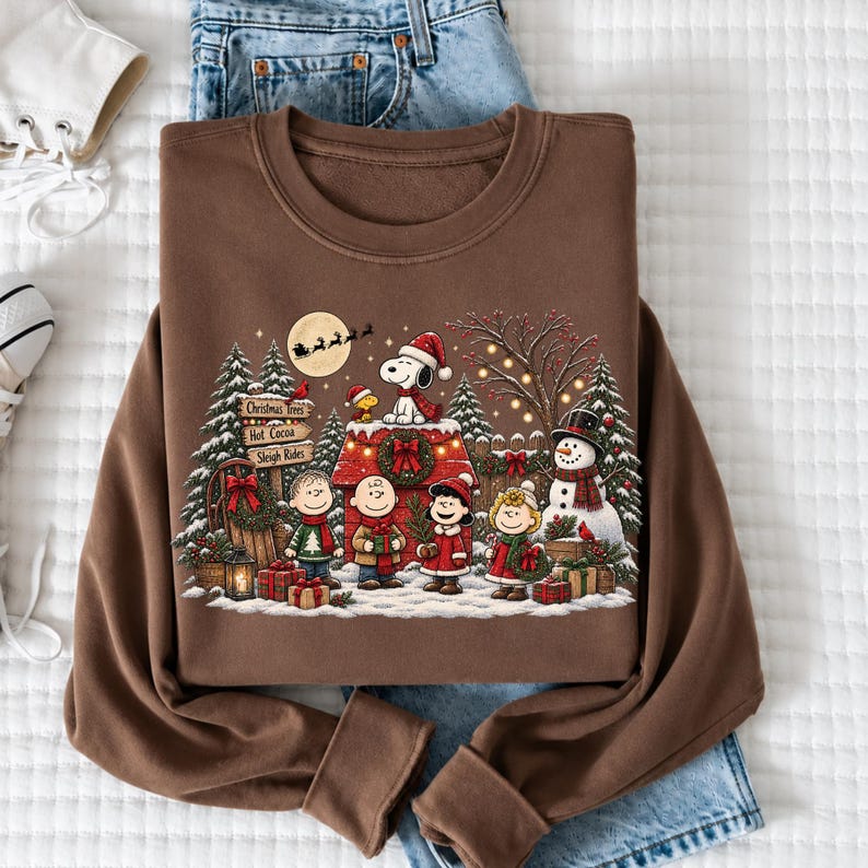 A brown sweatshirt with a Christmas-themed design featuring characters from the Peanuts comic strip, including Snoopy, Charlie Brown, and Woodstock, along with a snowy winter scene.