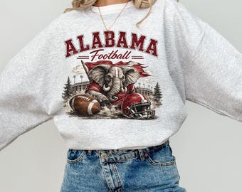 May include: A light grey crewneck sweatshirt featuring a graphic print with the text ALABAMA Football. The design includes an illustration of an elephant mascot, a brown football, and a red football helmet set against a stadium background.