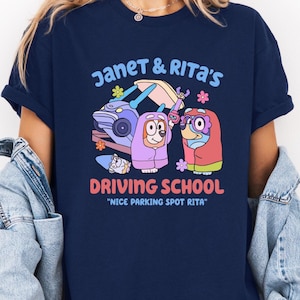 May include: Navy blue t-shirt featuring a colorful graphic with the text "Janet & Rita's Driving School." The design includes cartoon characters and a car. The phrase "Nice Parking Spot Rita" is also included.