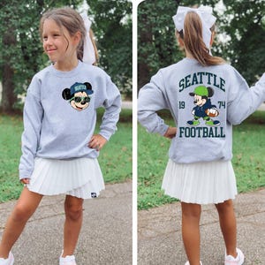 Puede incluir: Una sudadera gris con un dise&ntilde;o de Mickey Mouse con una gorra de b&eacute;isbol de Seattle y gafas de sol. La parte trasera de la sudadera muestra "SEATTLE FOOTBALL" con un jugador de f&uacute;tbol de Mickey Mouse y los n&uacute;meros "19" y "74". La sudadera se combina con una falda plisada blanca.