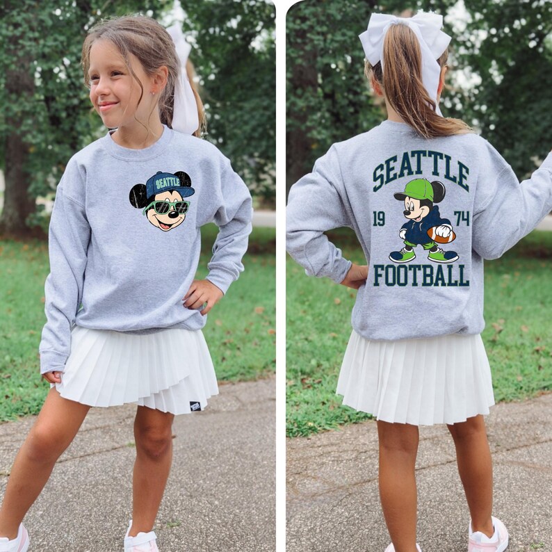 Puede incluir: Una sudadera gris con un dise&ntilde;o de Mickey Mouse con una gorra de b&eacute;isbol de Seattle y gafas de sol. La parte trasera de la sudadera muestra "SEATTLE FOOTBALL" con un jugador de f&uacute;tbol de Mickey Mouse y los n&uacute;meros "19" y "74". La sudadera se combina con una falda plisada blanca.