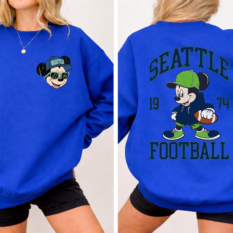 Puede incluir: Sudadera azul real con un rat&oacute;n de dibujos animados con gafas de sol y un sombrero con el texto "SEATTLE". La parte posterior de la sudadera muestra el texto "SEATTLE FOOTBALL" con un rat&oacute;n de dibujos animados sosteniendo un bal&oacute;n de f&uacute;tbol.
