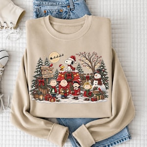 A beige sweatshirt with a Christmas-themed design featuring characters from the Peanuts cartoon, including Snoopy, Charlie Brown, and Woodstock, along with a snowy winter scene.