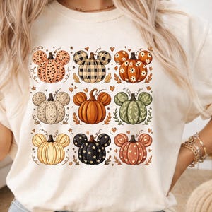 May include: A cream-colored t-shirt featuring a grid of nine pumpkin illustrations, each shaped like mouse ears. The pumpkins display various patterns including leopard print, black and white plaid, floral, polka dots, solid orange, and stars.