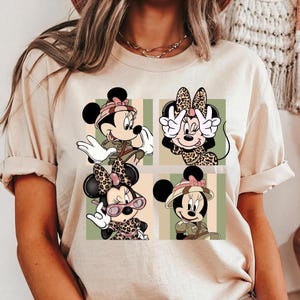 May include: A cream-colored short-sleeve t-shirt featuring a graphic print of four squares, each containing a different illustration of Minnie Mouse. The designs include Minnie in safari gear and leopard print outfits with pink accents against striped backgrounds.