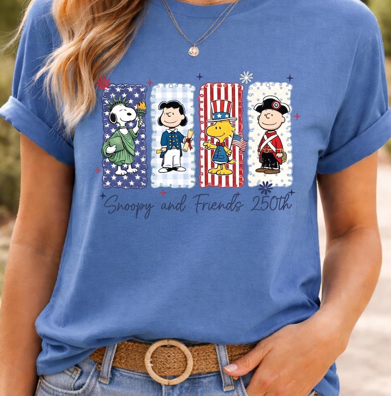 Puede incluir: Camiseta azul con un dise&ntilde;o de Snoopy y sus amigos con atuendos patri&oacute;ticos. El dise&ntilde;o incluye a Snoopy como la Estatua de la Libertad y otros personajes en rojo, blanco y azul. El texto "Snoopy and Friends 250th" est&aacute; impreso debajo.