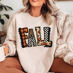 May include: A cream-colored crewneck sweatshirt featuring the word FALL in large block letters across the chest. Each letter has a unique pattern including orange plaid, leopard print, floral, and black and white checkerboard, outlined in black lace.