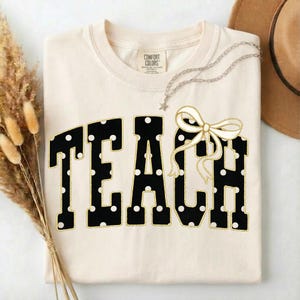 May include: A cream-colored short-sleeve t-shirt featuring the word TEACH in large black block letters with white polka dots and a gold outline. A decorative white ribbon bow is attached to the letter H. The shirt has a Comfort Colors brand label.