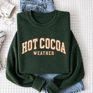 A green sweatshirt with the text "HOT COCOA WEATHER" printed on it, along with a pair of jeans and a white shoe.