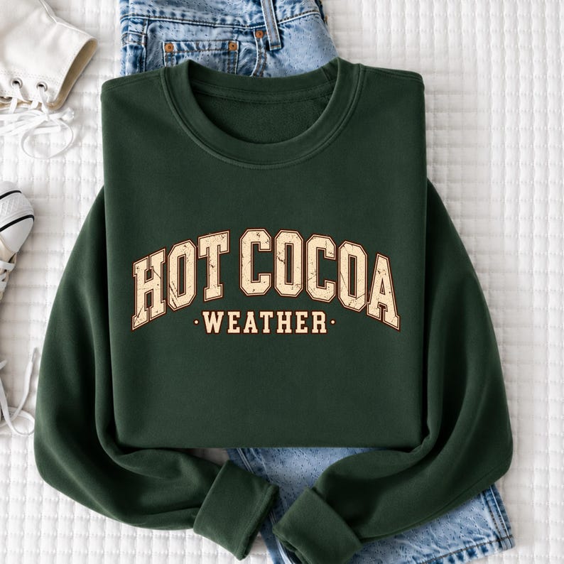 A green sweatshirt with the text "HOT COCOA WEATHER" printed on it, along with a pair of jeans and a white shoe.