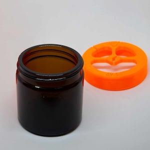 Può includere: Un barattolo di vetro marrone scuro con un'ampia apertura e un coperchio di plastica arancione brillante con un design a tre fori. Il coperchio ha la scritta "APS" stampata.