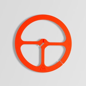 May include: Orange plastic ring with three spokes and a small triangle logo. The ring is designed to hold a pot or pan over a campfire.