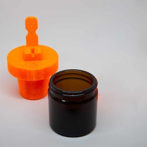 Può includere: Un barattolo di plastica arancione con un coperchio che ha una leva per aprirlo. Il barattolo è accanto a un barattolo di vetro marrone con un coperchio.