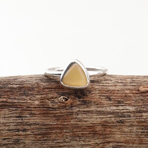 Könnte beinhalten: Ein silberner Ring mit einem dreieckigen, hellgelben Stein. Der Ring liegt auf einer Holzoberfläche, wobei das Design des Rings und die Farbe des Steins im Mittelpunkt stehen. Der Ring hat ein schmales, schlichtes Band.