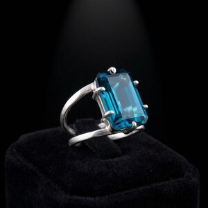 Könnte beinhalten: Ein silberner Ring mit einem großen, rechteckigen, teal-farbenen Edelstein. Der Edelstein ist in einer Krappenfassung gefasst, und das Band hat ein geteiltes Design. Der Ring wird auf einer schwarzen Oberfläche präsentiert, die von einem Scheinwerfer beleuchtet wird.