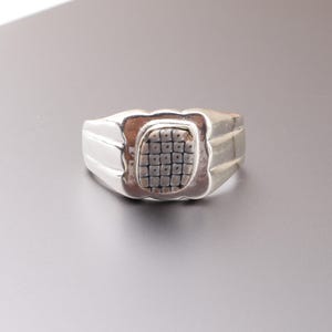 May include: A silver ring with a rectangular, textured center stone. The ring has a wide band with a ridged design on the sides. The center stone has a grid-like pattern.
