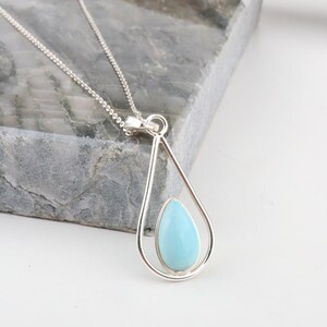 May include: A silver necklace featuring a teardrop-shaped pendant frame that holds a smaller, smooth, light blue stone in a matching teardrop shape. The pendant is suspended from a delicate silver chain with small, interconnected links.