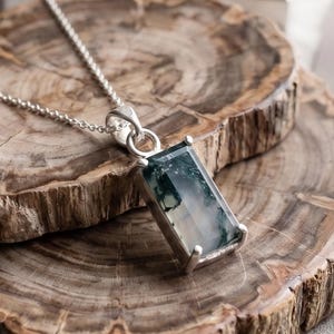 May include: A rectangular pendant necklace with a silver chain. The pendant features a green and white stone set in a silver frame. The necklace is displayed on a wooden surface with a natural grain pattern.