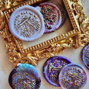 Pode incluir: Um conjunto de seis selos de cera com um design marmorizado roxo, azul e branco. Os selos apresentam um OVNI dourado com o texto "Anywhere But Here" ao redor da borda.