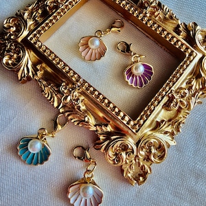 May include: Four gold-tone shell charms with pearl accents. The shells are pink, purple, teal, and white. The charms are displayed on a gold ornate frame.
