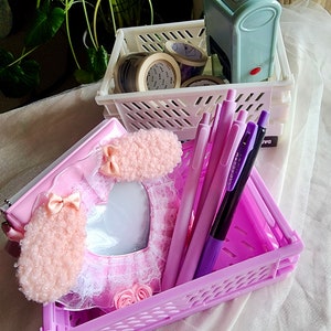 Puede incluir: Una caja de plástico rosa llena de bolígrafos y una libreta rosa en forma de corazón esponjosa con un borde de encaje. La caja está sobre una superficie blanca.