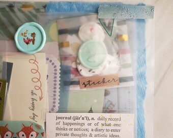 May include: A clear plastic envelope filled with colorful paper scraps, stickers, and washi tape. The envelope has a blue and green patterned design. The text "journal (jur'n'l), n. daily record of happenings or of what one thinks or notices; a diary to enter private thoughts & artistic ideas." is printed on a white label.