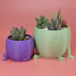 May include: Two small, colorful, 3D printed planters with legs, each holding a succulent plant. The planter on the left is purple and the planter on the right is green with a smiley face.