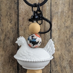 May include: White chicken-shaped keychain with a black clasp. The keychain features a white ceramic chicken, a floral patterned bead, and other beads in shades of tan and black. The background is a wooden surface.