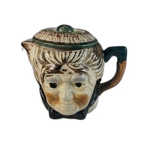 May include: A vintage ceramic teapot shaped like a man's face. The teapot features a cream-colored face with brown hair, a black bow tie, and a green handle. The lid is brown and green.