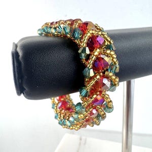 May include: A beaded bracelet featuring red, teal, and gold beads. The bracelet is displayed on a black stand. The beads are arranged in a pattern, with larger red faceted beads interspersed with smaller teal and gold beads.