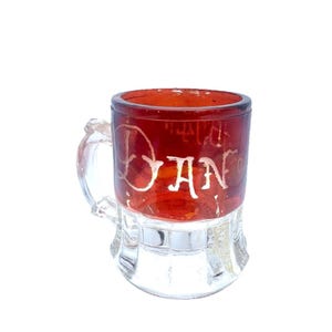 May include: A small, clear glass mug with a red upper portion and a clear base. The word "DAN" is written in white script on the red section. The mug has a small handle and a decorative base.