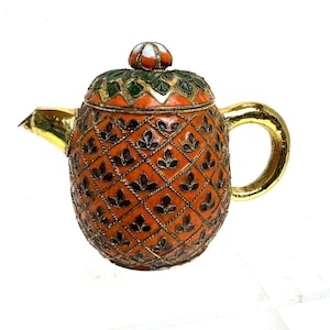 Puede incluir: Una tetera naranja y dorada ornamentada. La tetera presenta un asa y un pico dorados, con un patrón detallado de hojas y diamantes en el cuerpo. La tapa es verde y naranja, coronada con un pequeño pomo. Un artículo decorativo.