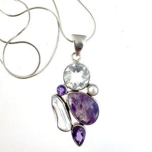 Könnte beinhalten: Eine silberne Anhängerkette mit einer Ansammlung von Edelsteinen. Der Anhänger enthält einen großen, klaren, runden Stein, einen großen lila Amethysten, einen kleinen lila Amethysten, eine weiße Perle und einen weißen, unregelmäßig geformten Stein.