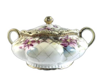 May include: A white porcelain sugar bowl with a lid, featuring a floral design with pink and purple flowers and gold accents. The bowl has a diamond pattern and a scalloped edge.