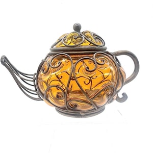 May include: A teapot featuring a translucent amber-colored glass body encased in a dark, weathered metal wire frame with intricate scrollwork patterns. The teapot includes a matching metal lid, a curved metal spout, and a rounded metal handle.